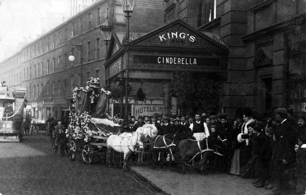 A horse-drawn carriage decorated with flowers on a city street. People are gathered around, and a theatre sign reads "King's Cinderella" above the entrance.