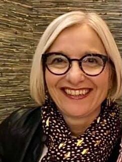 A woman with short blonde hair and round glasses smiles at the camera, wearing a dark jacket and a patterned scarf, with a textured background behind her.
