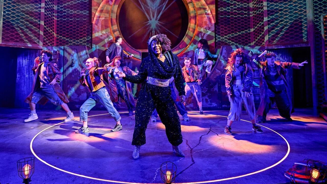 A group of performers in colorful costumes dance and pose energetically on a stage with vibrant lighting and geometric backdrops, bringing scenes from the Percy Jackson Musical to life.