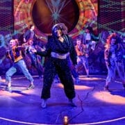 A group of performers in colorful costumes dance and pose energetically on a stage with vibrant lighting and geometric backdrops, bringing scenes from the Percy Jackson Musical to life.