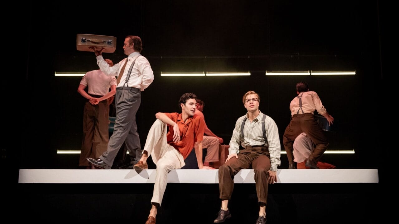 Seven people in vintage-style clothing perform on stage, evoking the allure of The Talented Mr. Ripley, with two seated on a raised platform as others stand or walk beneath sleek horizontal lights.