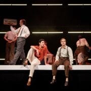 Seven people in vintage-style clothing perform on stage, evoking the allure of The Talented Mr. Ripley, with two seated on a raised platform as others stand or walk beneath sleek horizontal lights.