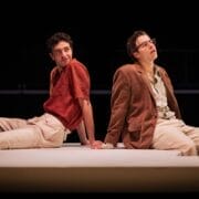 Two men sit back-to-back on a white platform, evoking The Talented Mr. Ripley; one wears a red shirt and beige pants, the other a brown jacket and striped shirt, with a glass nearby.