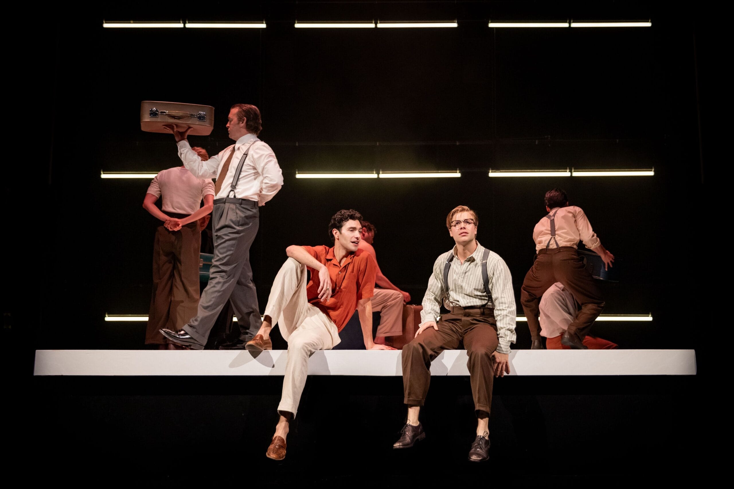 Seven people in vintage-style clothing perform on stage, evoking the allure of The Talented Mr. Ripley, with two seated on a raised platform as others stand or walk beneath sleek horizontal lights.