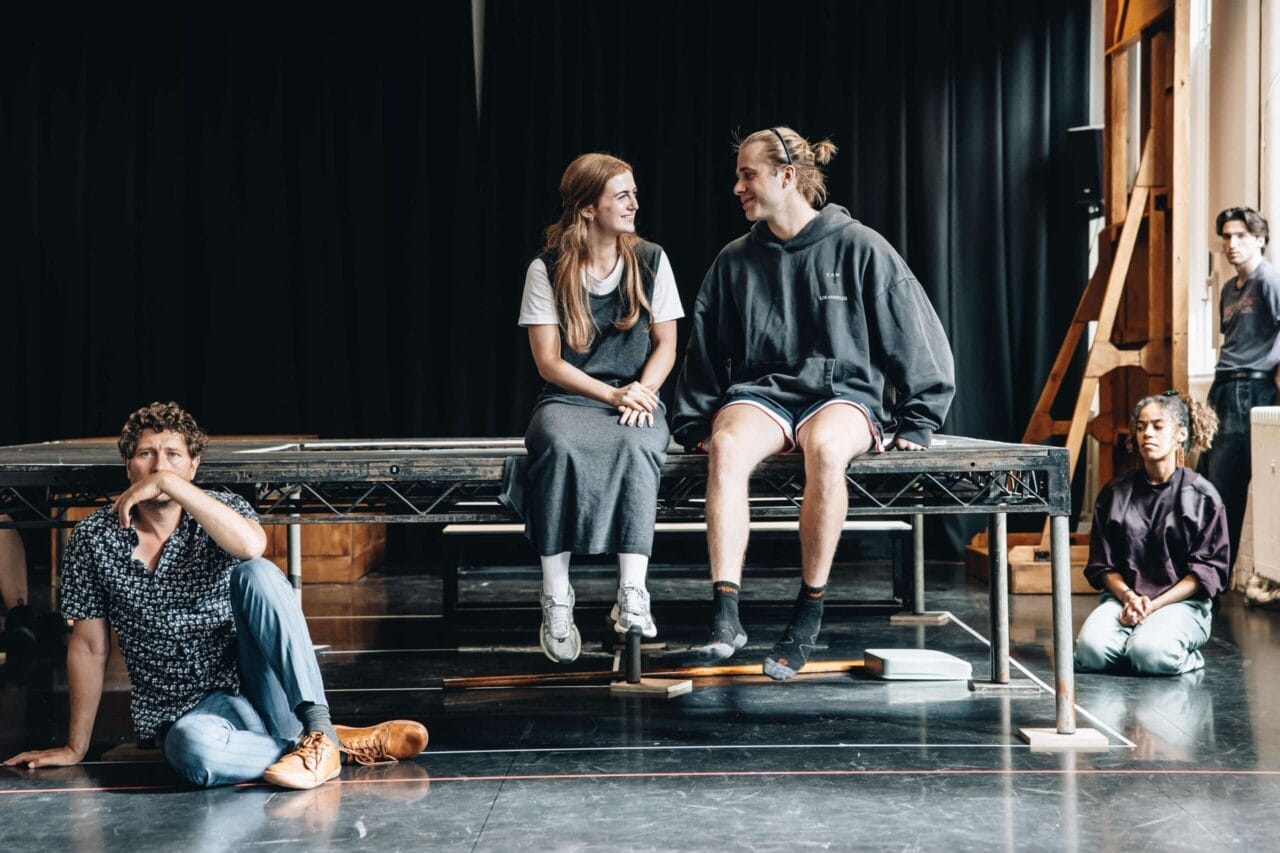 Four people are in a rehearsal space; two sit on a platform talking, one sits on the floor thinking, and another kneels near the edge of the stage, echoing the tense atmosphere of The Talented Mr. Ripley.