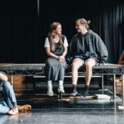 Four people are in a rehearsal space; two sit on a platform talking, one sits on the floor thinking, and another kneels near the edge of the stage, echoing the tense atmosphere of The Talented Mr. Ripley.