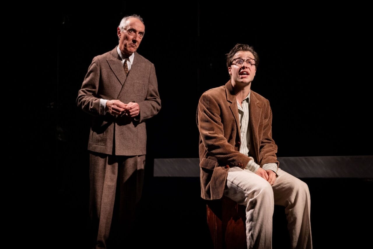 Two men in brown suits are on stage under dramatic lighting—one stands with hands clasped, while the other, reminiscent of The Talented Mr. Ripley, sits on a box, appearing distressed.