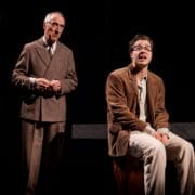 Two men in brown suits are on stage under dramatic lighting—one stands with hands clasped, while the other, reminiscent of The Talented Mr. Ripley, sits on a box, appearing distressed.
