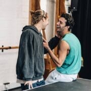 Two young men smiling and laughing together indoors, one standing and the other sitting on the edge of a table in a casual, light-filled room, echoing the camaraderie seen in The Talented Mr. Ripley.