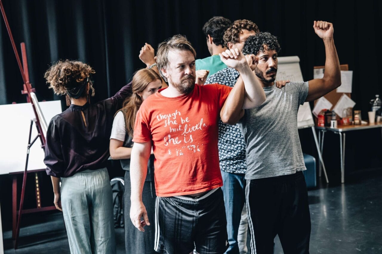 A group of people stand in close formation indoors, some raising their arms, appearing to rehearse or perform a scene inspired by The Talented Mr. Ripley.