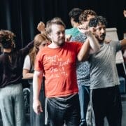 A group of people stand in close formation indoors, some raising their arms, appearing to rehearse or perform a scene inspired by The Talented Mr. Ripley.