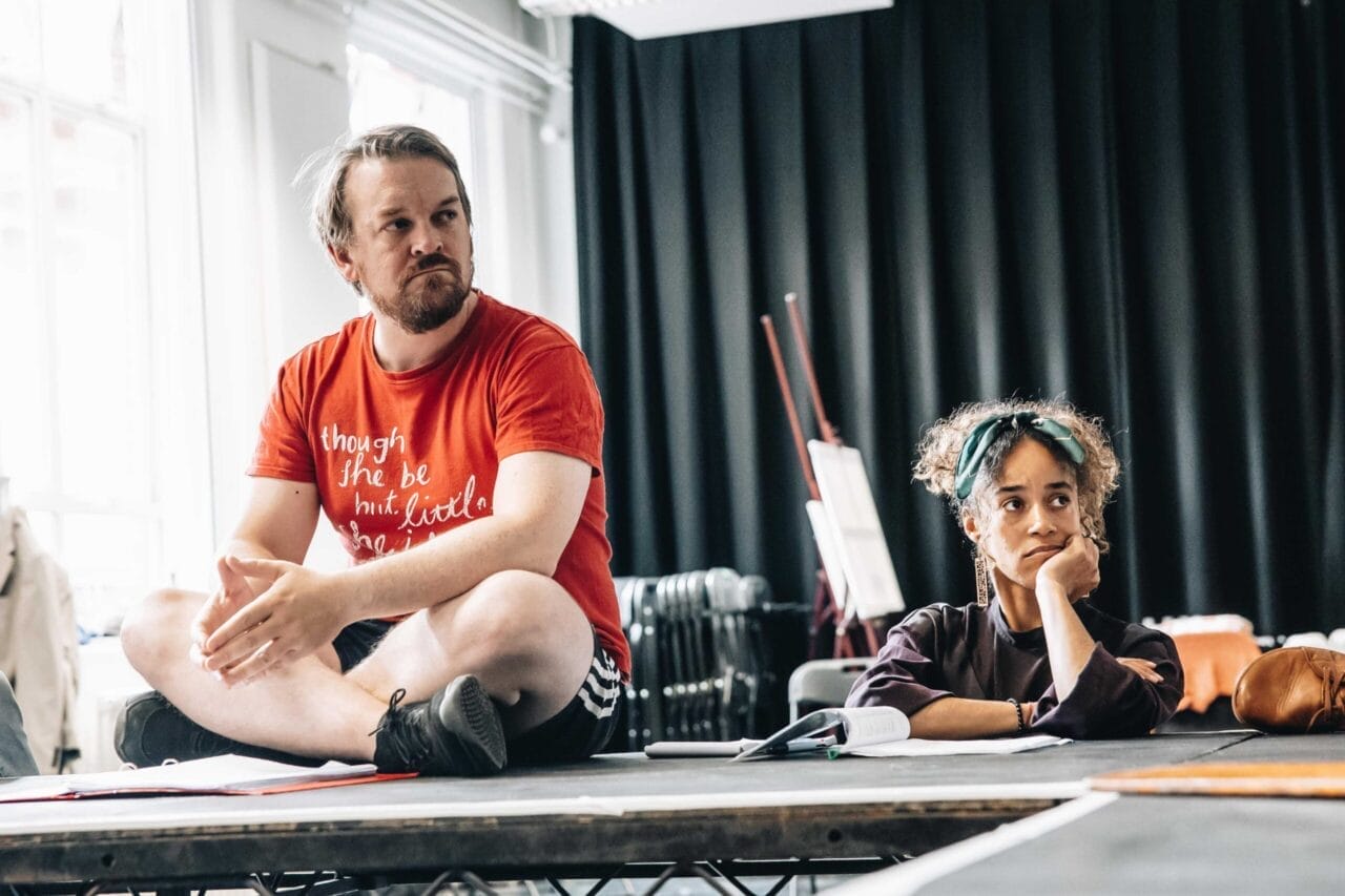 A man in a red T-shirt sits cross-legged on a table, while a woman beside him rests her chin on her hand. They appear to be in a discussion or rehearsal space with black curtains, evoking the atmosphere of a Talented Mr. Ripley audition.