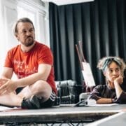 A man in a red T-shirt sits cross-legged on a table, while a woman beside him rests her chin on her hand. They appear to be in a discussion or rehearsal space with black curtains, evoking the atmosphere of a Talented Mr. Ripley audition.