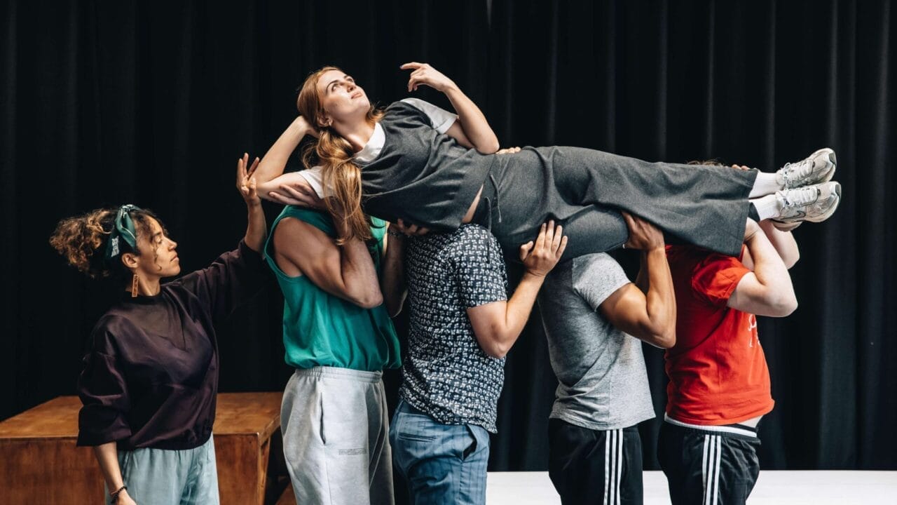 A group of people holds a woman horizontally above their heads, reminiscent of a dramatic scene from The Talented Mr. Ripley; she is wearing gray clothes and looking upward, while others support her in front of a black curtain backdrop.