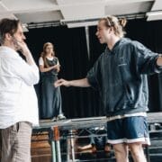 Three people rehearse on stage; one man gestures with open arms while talking to another, as a woman in the background watches them—capturing tension reminiscent of The Talented Mr. Ripley.