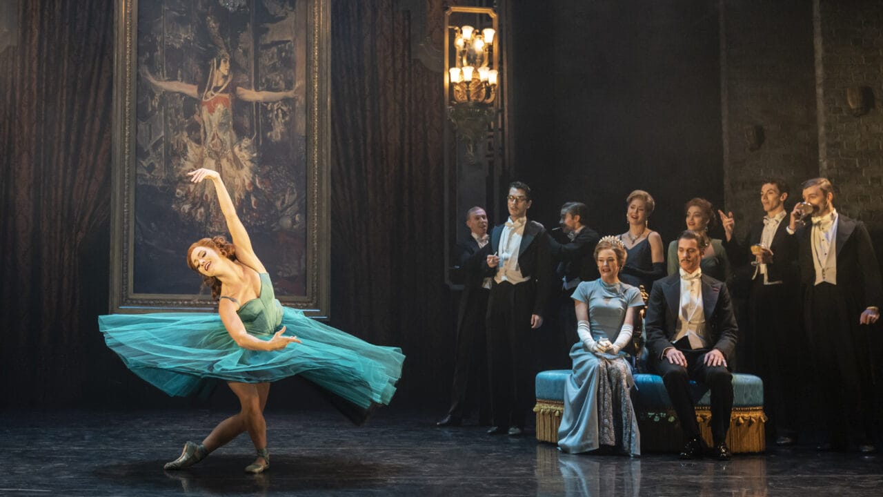 A ballerina in a blue dress performs on stage in front of a painting, while formally dressed people watch from the side and a chandelier hangs overhead.
