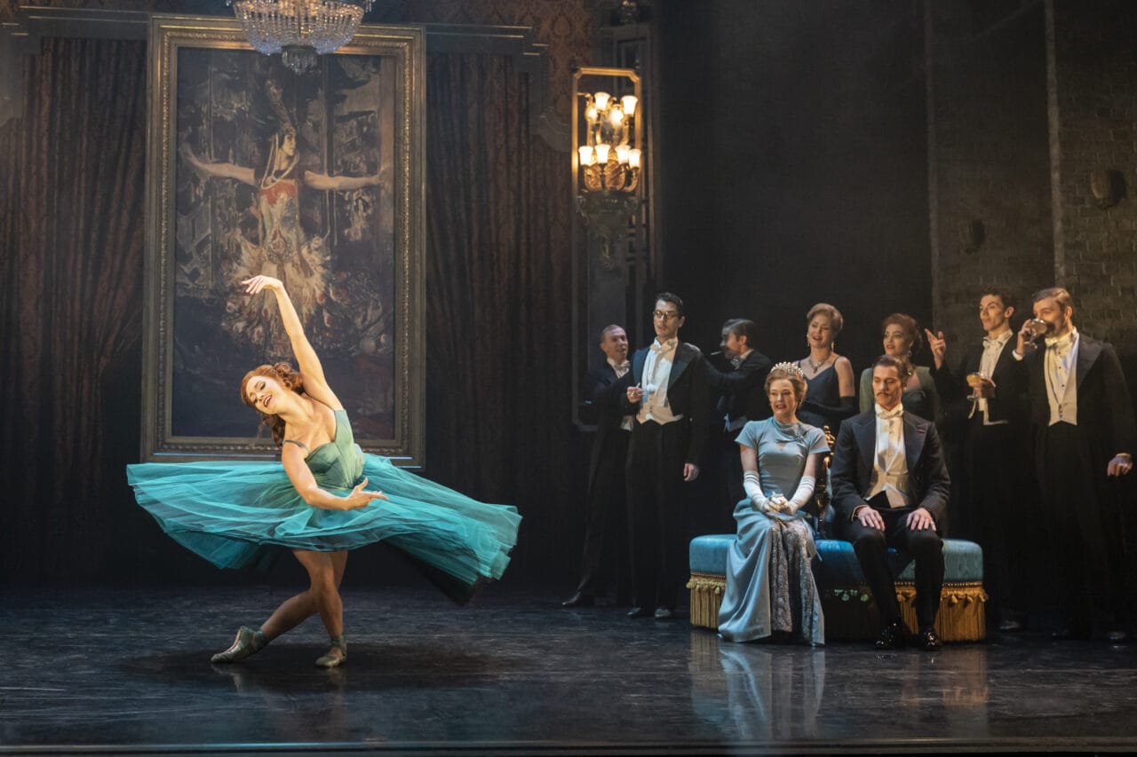 A ballerina in a blue dress performs on stage in front of a painting, while formally dressed people watch from the side and a chandelier hangs overhead.