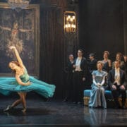 A ballerina in a blue dress performs on stage in front of a painting, while formally dressed people watch from the side and a chandelier hangs overhead.