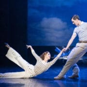 A woman in white pants and a striped shirt poses on the floor, holding hands with a standing man in similar attire, performing on stage with a seascape backdrop.