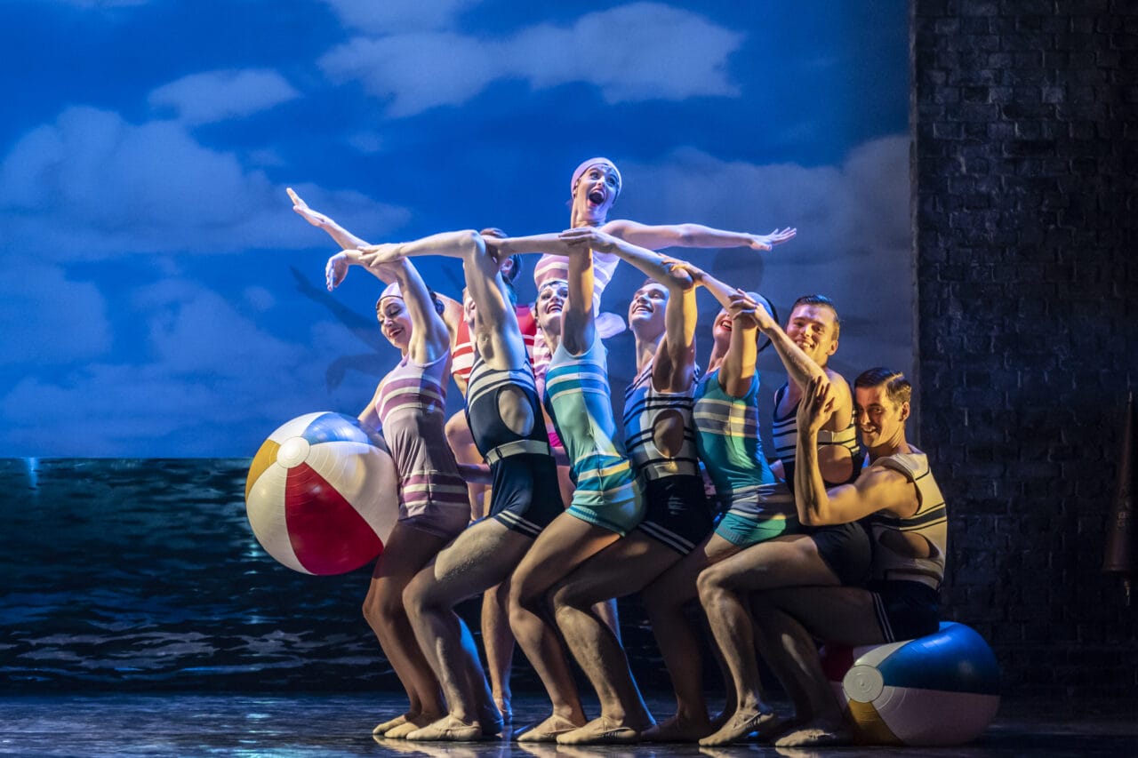 A group of dancers in vintage swimsuits pose together on stage with beach balls, against a backdrop of blue sky and ocean.