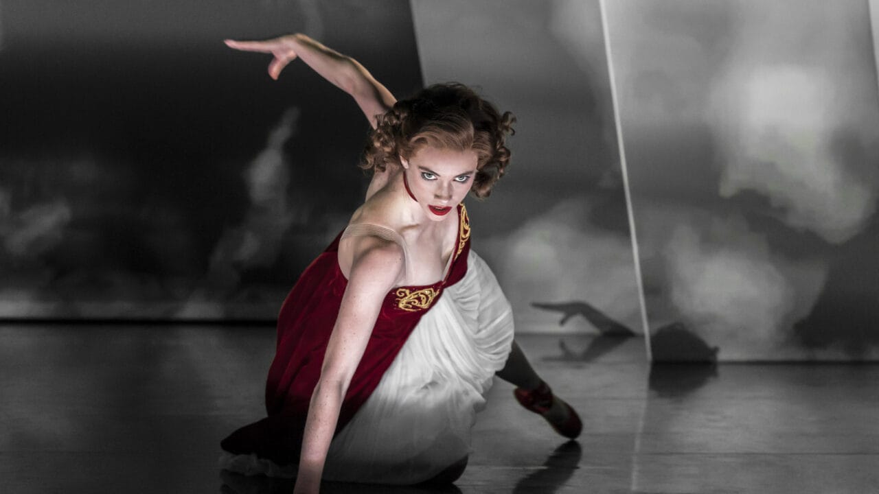 A dancer in a red and white dress poses close to the floor with one arm extended, set against a dramatic, moody background.