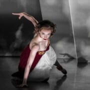 A dancer in a red and white dress poses close to the floor with one arm extended, set against a dramatic, moody background.