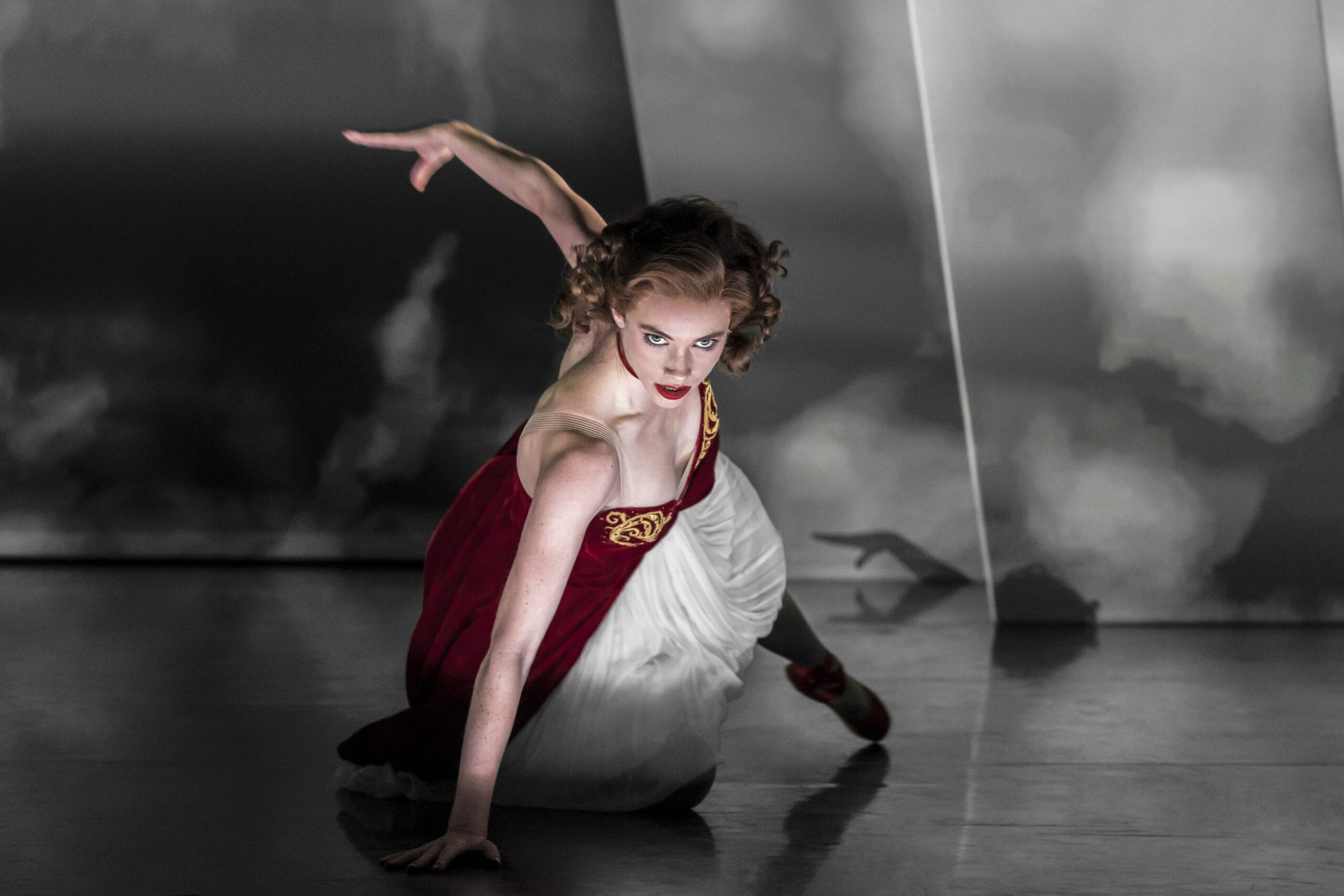 A dancer in a red and white dress poses close to the floor with one arm extended, set against a dramatic, moody background.