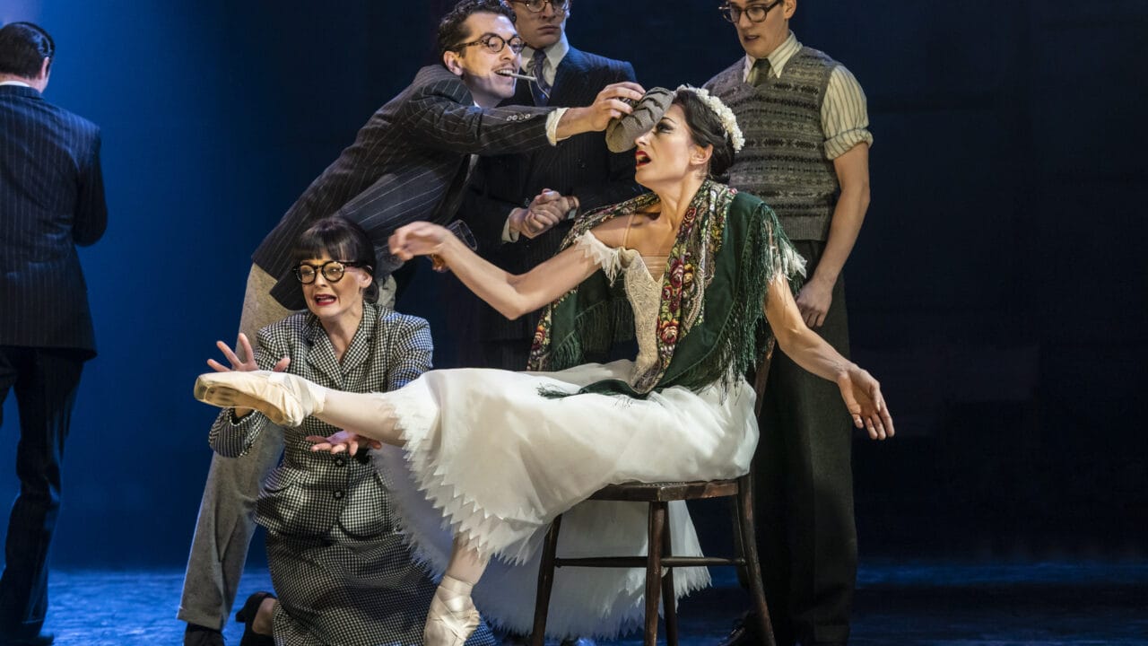A group of actors in vintage costumes surround a seated ballerina on stage; one man reaches toward her head as she balances en pointe on one foot.