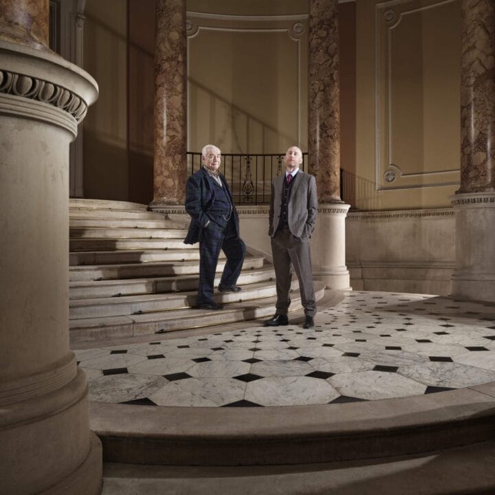 Two men in suits stand on a marble staircase in an ornate interior with tall columns and patterned floor tiles, ready to make it happen.