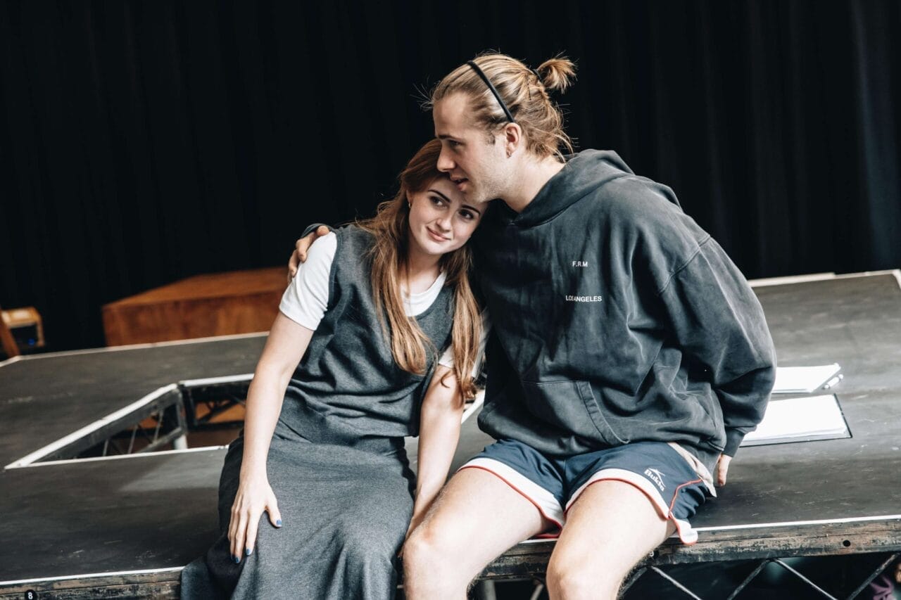 Two people sit closely on a stage, one resting their head on the other's. They appear relaxed and affectionate, with scripts beside them—evoking an intimate moment reminiscent of The Talented Mr. Ripley movie.