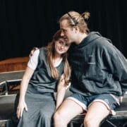 Two people sit closely on a stage, one resting their head on the other's. They appear relaxed and affectionate, with scripts beside them—evoking an intimate moment reminiscent of The Talented Mr. Ripley movie.