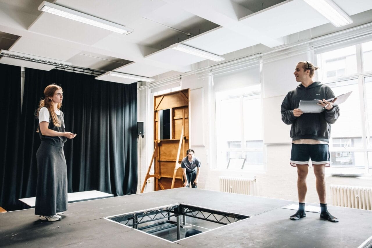 Three people in a rehearsal room: two stand on a stage holding scripts from The Talented Mr. Ripley, while one person crouches in the background near a ladder.