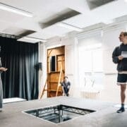 Three people in a rehearsal room: two stand on a stage holding scripts from The Talented Mr. Ripley, while one person crouches in the background near a ladder.