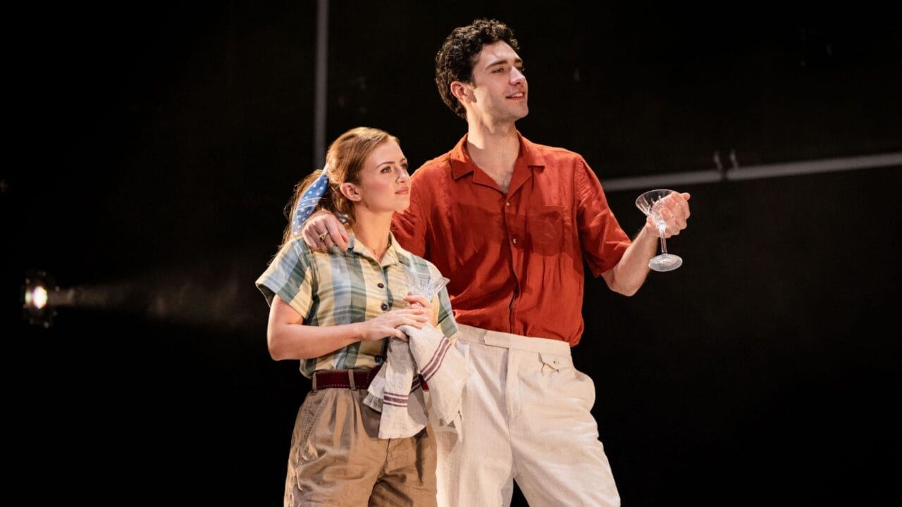 A woman in casual clothes holds a dish towel while a man in a red shirt and white pants stands beside her holding a cocktail glass, evoking the stylish flair of The Talented Mr. Ripley as they both take the stage.