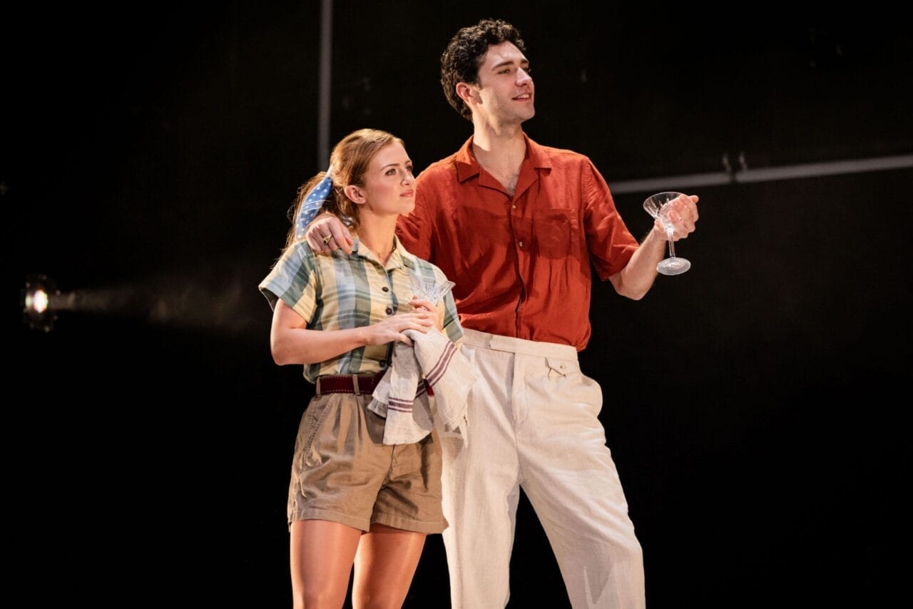 A woman in casual clothes holds a dish towel while a man in a red shirt and white pants stands beside her holding a cocktail glass, evoking the stylish flair of The Talented Mr. Ripley as they both take the stage.