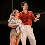 A woman in casual clothes holds a dish towel while a man in a red shirt and white pants stands beside her holding a cocktail glass, evoking the stylish flair of The Talented Mr. Ripley as they both take the stage.