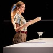 A woman stands at a white table holding an open book, with a martini glass and a piece of paper on the table, evoking the classic elegance of The Talented Mr. Ripley. She wears a plaid shirt, brown pants, and a blue hair ribbon.