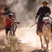 A man rides a white horse on a dusty trail, embodying the adventurous spirit of the Red Programme, while another person pushes a mountain bike uphill behind him.