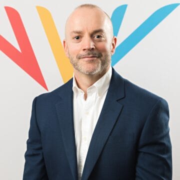A man in a navy suit and white shirt poses in front of a white background with red, yellow, and blue V-shaped graphics.