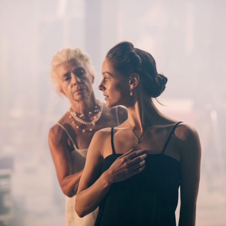 Two women stand in a hazy setting. A woman in a white dress gestures towards a younger woman in black, who looks forward.