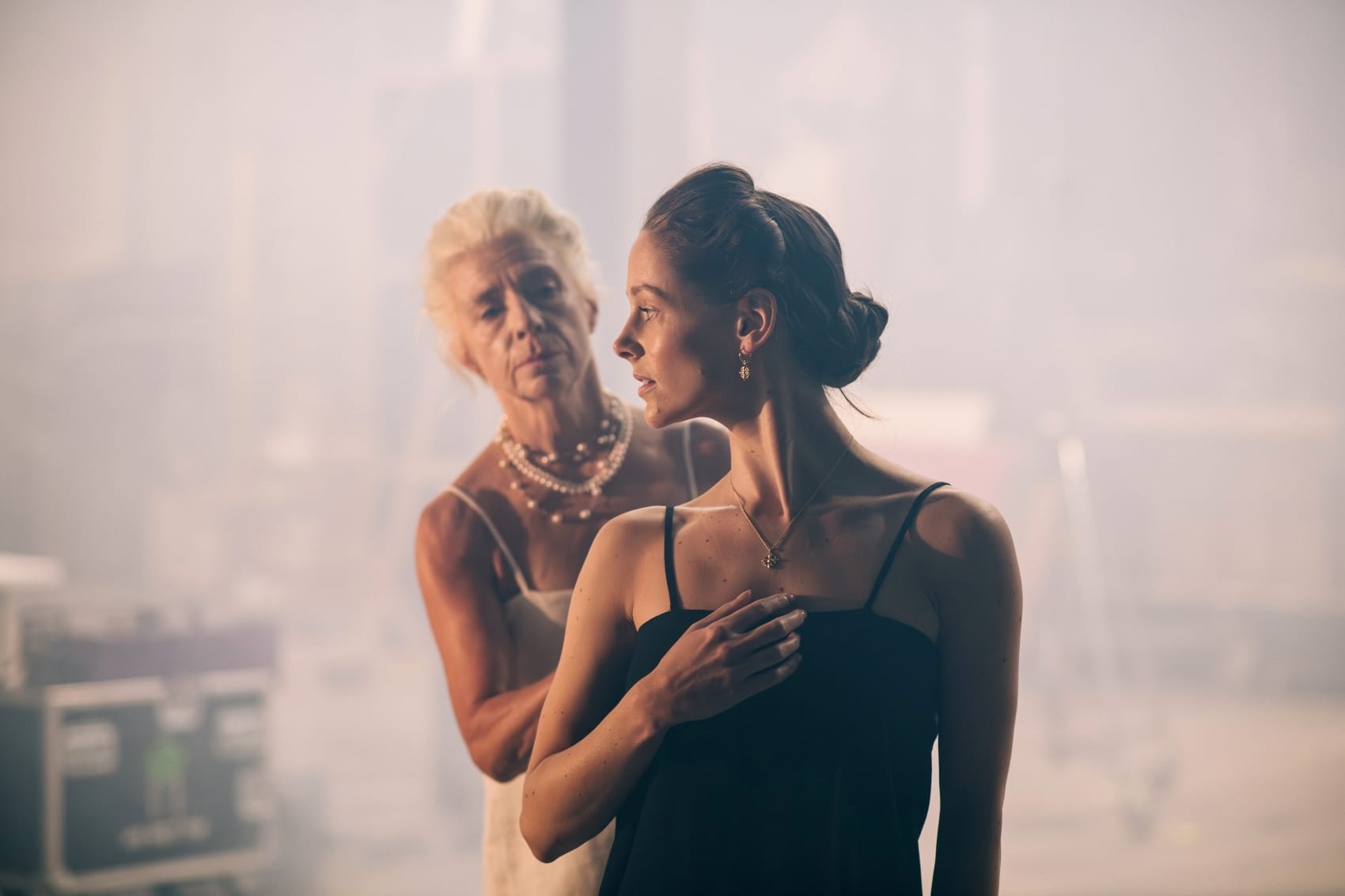 Two women stand in a hazy setting. A woman in a white dress gestures towards a younger woman in black, who looks forward.