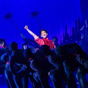 A group of performers dressed as chimney sweeps dance onstage, with one person in a red shirt at the center raising their fist under blue stage lighting.