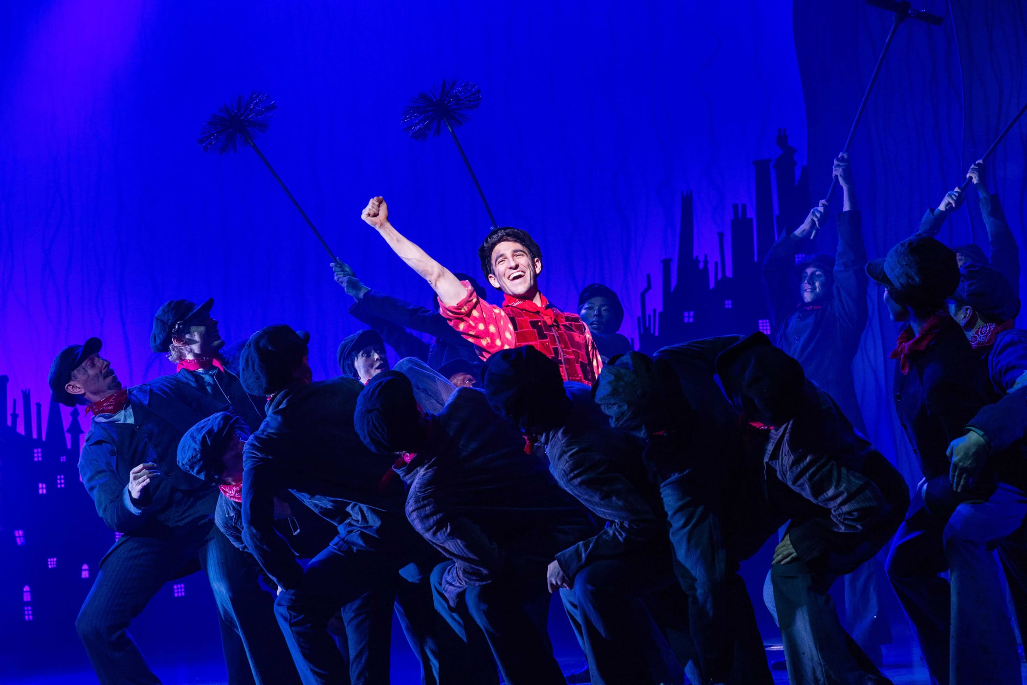 A group of performers dressed as chimney sweeps dance onstage, with one person in a red shirt at the center raising their fist under blue stage lighting.