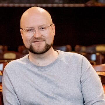 A person with glasses and a beard, wearing a light gray sweatshirt, sits and smiles in a theatre with ornate chairs in the background.