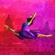 A dancer in a purple dress performs a split leap against a vibrant pink and red background with city buildings and domes.