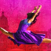Ballet dancer in a purple dress performs a split leap against a vibrant pink and red cityscape background.