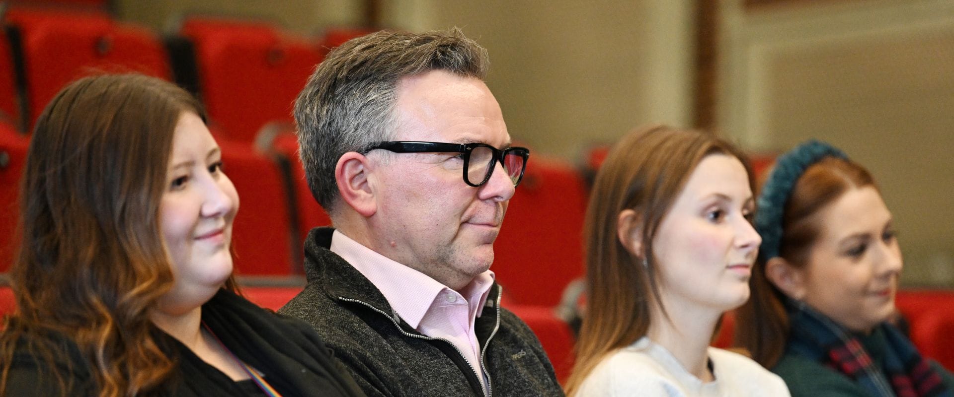 Four individuals seated in red auditorium chairs, attentively facing forward.