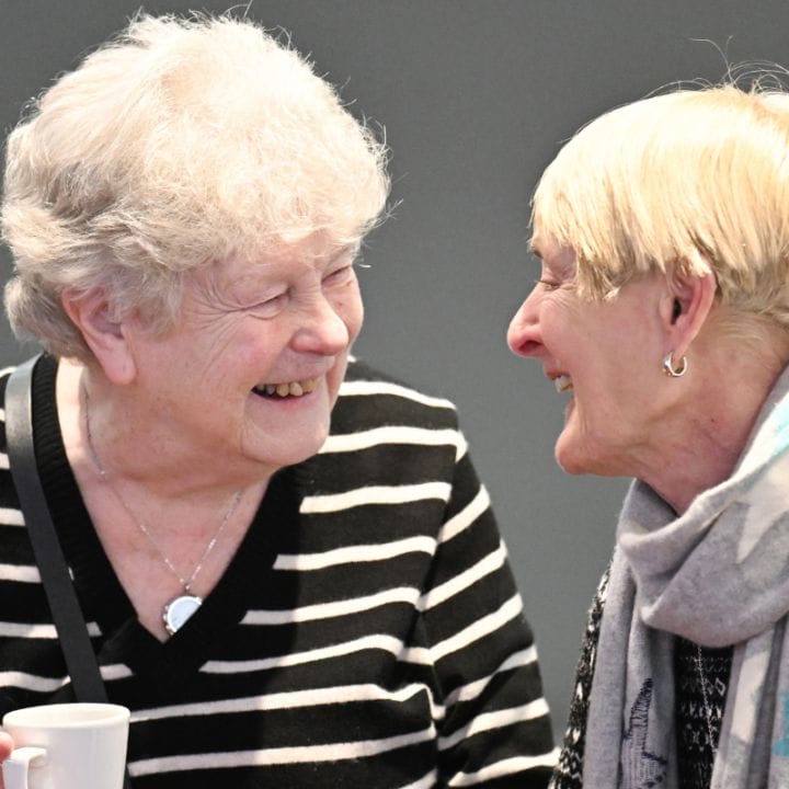 Two elderly women smiling at each other, one holding a cup. Both are wearing striped sweaters and appear to be engaged in conversation.