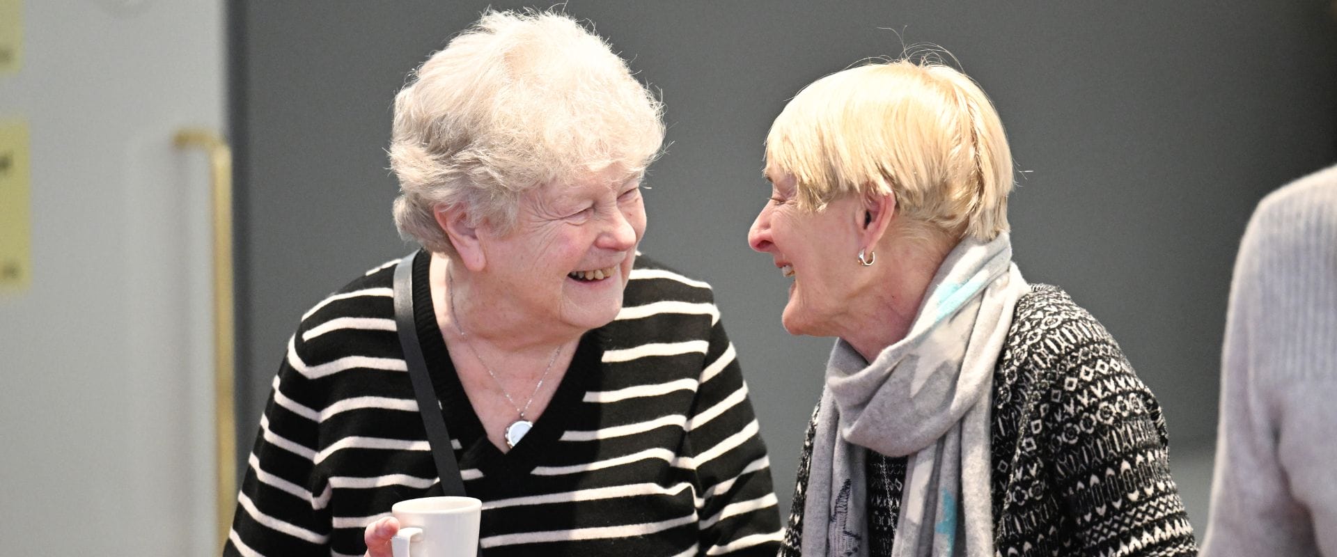 Two elderly women smiling at each other, one holding a cup. Both are wearing striped sweaters and appear to be engaged in conversation.