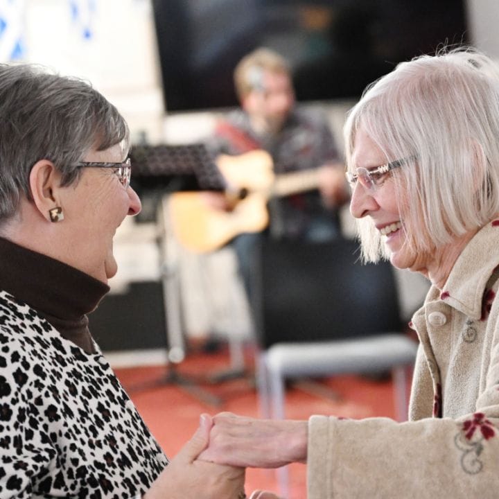 Two elderly women are dancing and smiling indoors. In the background, musicians are playing instruments. Scottish flags are visible in the decor.